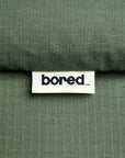 Close-up of a textured olive green 100% cotton ripstop fabric with a white sewn-in label reading 'bored'.