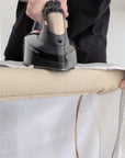 A close-up of a person using a handheld steam iron on a beige ironing board cover, with a white shirt draped over.