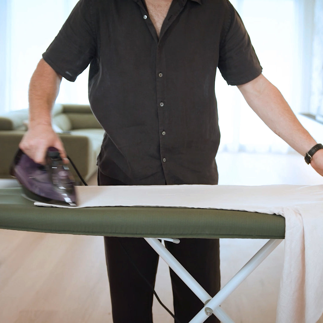 A man ironing linen trousers on an olive green ironing board cover in a modern interior.