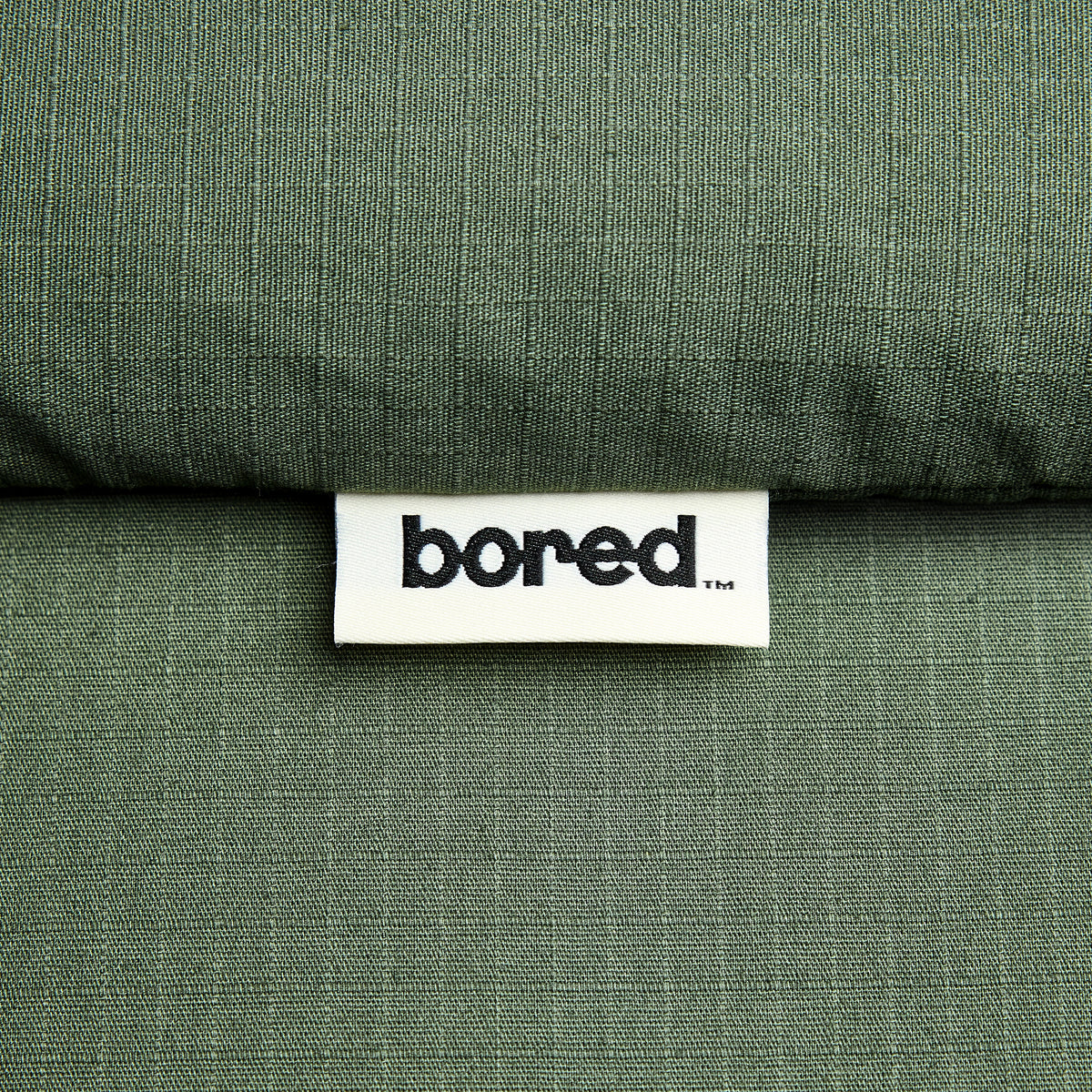Close-up of a textured olive green 100% cotton ripstop fabric with a white sewn-in label reading 'bored'.