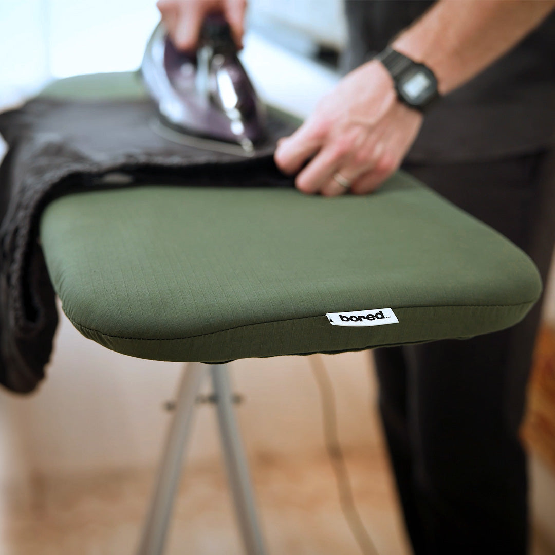An olive green ironing board cover with a small 'bored' brand tag, being ironed by a person in a home setting.