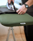 An olive green ironing board cover with a small 'bored' brand tag, being ironed by a person in a home setting.