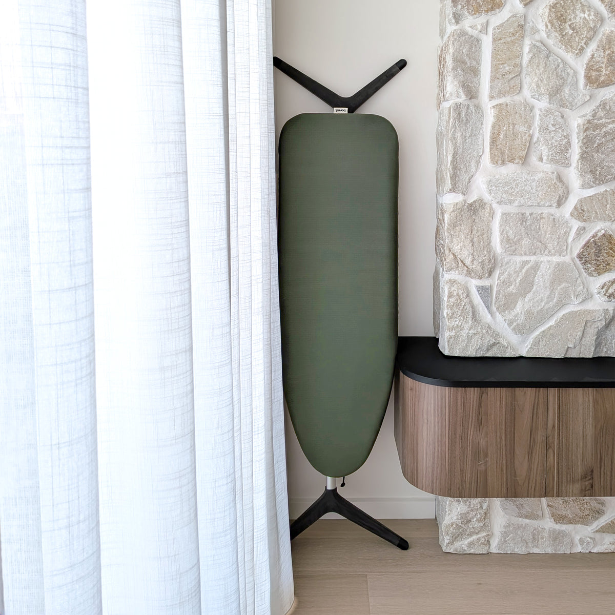 An ironing board with an olive green cover, positioned next to a white curtain and a stone fireplace.