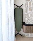 An ironing board with an olive green cover, positioned next to a white curtain and a stone fireplace.